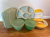 Photo showing front perspective of the vintage Tupperware pieces laid on wooden floor: two mint green mixing bowls with lids, five-compartment divided tray, two rectangular containers, and yellow handled container.