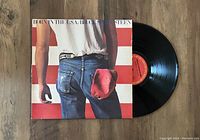 Photo showing the vinyl record LP partially out of its original album jacket featuring the iconic cover art of Bruce Springsteen's Born In The USA album with his back turned, American flag stripes background, and red cap in hand.