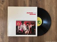 Vinyl record partially out of white jacket with band photo and red text, shot on wooden background.