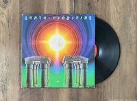 Photo showing the Earth Wind and Fire 'I Am' album jacket and the vinyl record partially removed from the sleeve on a wooden surface. Album cover features a radiant sun/starburst design and ancient pillars.