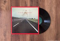 Vinyl LP partially pulled out of the album cover, showing green label on the record and album cover with a road, sky and red border.