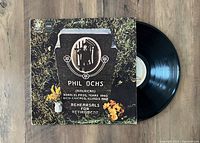Photo of Phil Ochs vinyl LP record and cover titled 'Rehearsals For Retirement'. Cover features a tombstone design with flowers, showing very good plus condition. Vinyl record visible alongside.