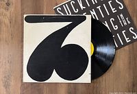 Vinyl record partially outside its cover showing front artwork with large black stylized '3' on white background, record has yellow label visible. Insert with text is partially visible behind the cover.