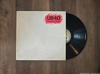 Photo of UB40 Present Arms vinyl LP record partly out of its original plain white jacket with red and black logo visible, placed on wooden surface.