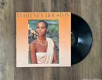 Color photo showing the front cover of Whitney Houston 1985 LP jacket and vinyl partially out of sleeve on wooden background