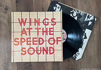 The Wings At The Speed Of Sound album jacket sleeve with visible red text and grid pattern, partially opened showing the black vinyl record and the black and white inner sleeve insert with band member photos.