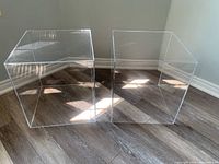 Two clear cube plexiglass tables side by side on wood floor with sunlight reflections.