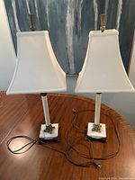 Front view of the two white table lamps on a wooden table, showing squared white marble bases, brass accents, and white fabric shades with cords visible.