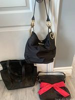 Photo showing all three handbags together: large black DE tote, black rope bag with pink bow, and small black fabric bag with gold accents.