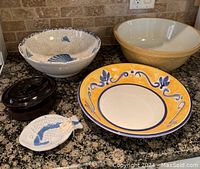 Five assorted bowls and dishes arranged on granite countertop: large beige bowl with embossed pattern, large white bowl with seashell pattern inside, large yellow bowl with blue floral design, mini black dish with lid, and white fish-shaped ladle dish with blue scales.