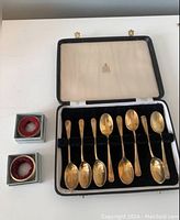 Set of 8 gold-tone small spoons arranged in original black presentation box with Birks logo. Two small round wine collars in separate boxes.