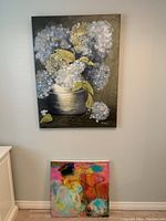 Large floral canvas art hanging on wall above smaller abstract canvas leaning on floor.