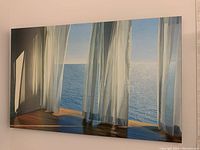 Large horizontal canvas mounted on a beige wall showing sheer white drapes flowing in front of a calm ocean scene under daylight.