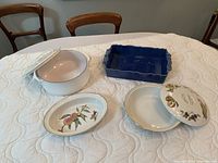 Four assorted ceramic dishes arranged on a table; white Dansk pot with lid, blue rectangular dish, and two oval dishes with painted fruit designs.