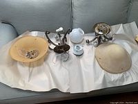 Two matching ceiling lights disassembled and laid out, showing beige glass shades, metal bases, wiring and mounting hardware on white paper on grey background.