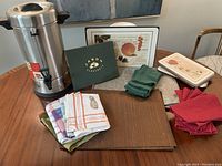 Large stainless steel coffee pot with black handles and lid, still in original box displayed on wood table with napkins and placemats