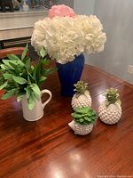 Photo of all decor items on wooden table including large blue vase with white flowers and pink flower, white pitcher with greenery, two white ceramic pineapples with green tops, and small white porcupine with green succulent.
