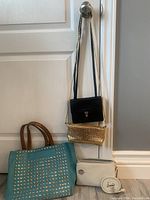 Photo showing four purses hanging on a knob: black Derek Alexander crossbody, gold sequin crossbody, off-white crossbody with strap, and turquoise studded handbag with brown handles
