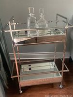 Front view of chrome-finished bar cart with two mirrored shelves and two square glass decanters on wooden floor against gray wall