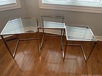 Three glass nesting tables shown together, displaying varying sizes with metallic frames and clear glass tops on wooden floor.