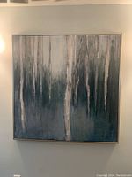 Front view of large square canvas painting of misty forest with vertical tree trunks in muted blues, greens, whites, and grays. Framed in brushed silver frame and hung on wall.