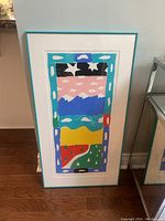 Full view of framed artwork showing colorful abstract landscape with turquoise frame on wooden floor.