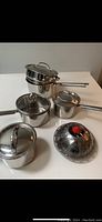 Photo showing four small stainless steel pots with lids and handles arranged, with one stainless steel steamer insert with a red handle.