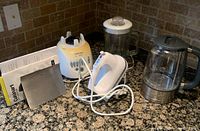 Photo showing a Black & Decker blender, Cuisinart electric kettle, and white hand mixer on a kitchen counter with granite finish and tiled backsplash. All appliances show signs of use and staining.