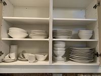 Cabinet with assorted white ceramic dishes including stacks of plates, bowls, mugs, sauce boats, and gravy boat