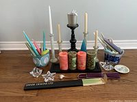 Wide view of lot showing several tall thin candles in holders, four large scented pillar candles on a black base, incense sticks in a box, glass votive holders, and two ceramic flower pots.