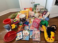 Wide shot showing assortment of kids toys, books, DVDs, stuffed toys including green stuffed alligator, red bucket, toy drum, toy saxophone, and multiple books and DVDs.