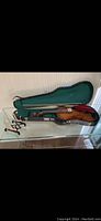 Violin with bow and red velvet cloth inside open hard case with green lining and wooden violin stand on glass table.