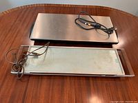 Both warming trays placed on wooden table: Salton brand brushed metal tray with black handles, Toastess branded glass top retro tray