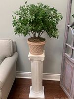 Full view of white Greek-style column pedestal with beige ceramic pot and artificial green plant on it, next to a sofa and window.