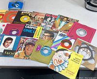 Photo of various 45 RPM vinyl singles with colorful and vintage sleeves, featuring artists and music titles as described by the seller.