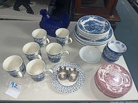 Overview of dinnerware set including mugs, plates, and small bowls in blue and white patterns with the blue glass rooster candy dish and two metal crown napkin rings.
