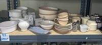 Shelf display of mixed cream and white serving ware including mixing bowls, ramekins, cups with saucers, creamer, pie bakers, and utensils.