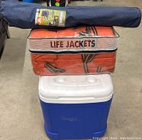 Stacked cooler, life jackets in storage bag, and folded chair on top, showing cooler bottom and bag labeling