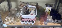 Photo showing shelves with kitchenware including stacked plates, egg cups, multiple colored drinking glasses, clear smaller glasses, blue mugs, and cutlery trays with kitchen tools inside.