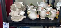 Shelf display showing Lenox cream and gold dishes on left, Goebel tulip design teapot set on right including creamer, sugar bowl, cups, small vase, and teapot with lid.