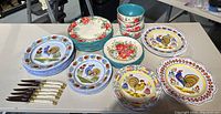 Full view of all plates, bowls, and knives arranged on table showing floral and rooster designs with varied colors.