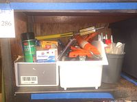 Shelf holding white bins filled with various adhesive tubes and bottles, some loose tools including caulking guns and a spray adhesive can