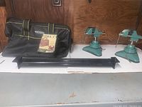 Photo shows two teal articulating arm vises with triangular rubberized bases, an 18-inch black metal bending brake, and a black tool bag with product tag. Items placed on white surface against wooden background.