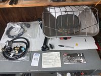 Photo showing the propane hose assembly including gas gauge and brass fittings, mounting post parts, and heater with protective wire guard, all arranged on a table.