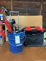 Overview showing blue bucket cart with telescoping handle and wheels filled with assorted hammers, measuring wheel propped on bucket rim, and Husky seat to the right