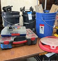 View of three plastic buckets—two black with wheeled handles and one blue containing metal trowels—plus two stacked clear plastic hardware cases and a round compartmentalized container on a table.