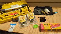 Wide photo of Alton laser level kit contents including yellow tripod, laser level device, two packaged laser markers, magnetic puck level, a small black pouch, brass plumb bob with string and safety goggles all on wooden surface