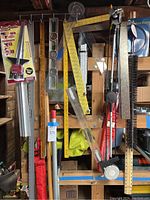 Wide view showing assorted measuring tools including yellow yard stick, clear plastic squares, levels, saw guide box, and scrapers hung on a wooden pegboard.