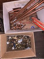 Two cardboard boxes filled with various brass, copper fittings and valves, copper pipes, pipe cutter tool, and Teflon tape visible in the images.