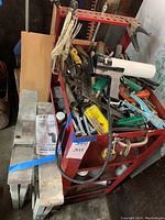 Photo showing top and side view of red steel utility cart loaded with assorted hand tools such as clamps, saws, and lubricants, plus folded metal saw horses strapped with blue tape leaning against the cart.